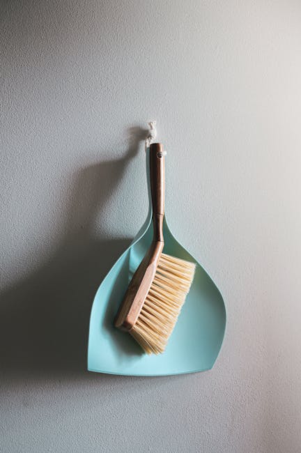 A light blue dustpan and a wooden-handled brush with beige bristles hanging on a white wall in a domestic setting, representing surface cleaning tools. The image highlights the cleanliness and organiz