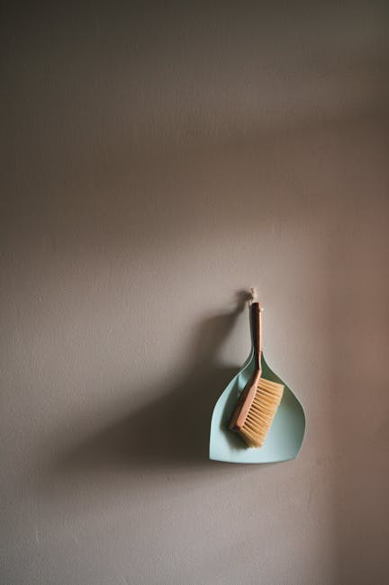 A light blue dustpan and a wooden-handled brush with beige bristles hanging on a white wall in a domestic setting, representing surface cleaning tools. The image highlights the cleanliness and organiz