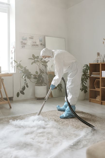 A person in a beige jumpsuit is engaged in surface cleaning, vacuuming a carpeted area within an indoor space. They are bent over, operating a yellow and black industrial vacuum cleaner with a long ho
